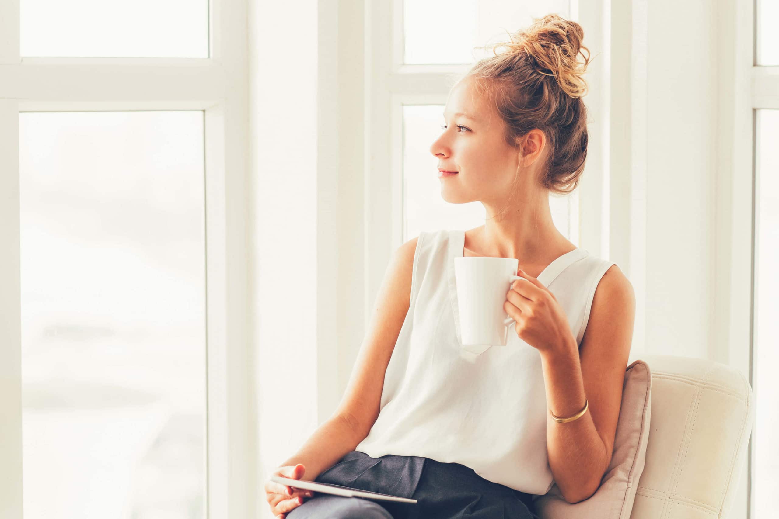 smiling woman sitting on cozy chair with mug near large windows